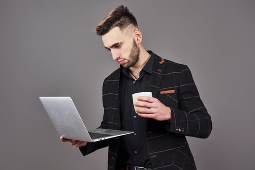 Busy bearded man in business clothes talking by smartphone and using laptop computer while holding cup of coffee in hand over gray background photo