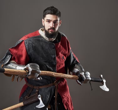 Medieval knight on grey background. Portrait of brutal dirty face warrior with chain mail armour red and black clothes and battle axe photo