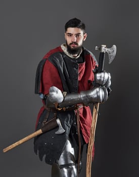 Medieval knight on grey background. Portrait of brutal dirty face warrior with chain mail armour red and black clothes and battle axe photo