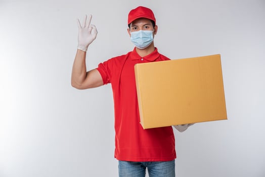 Image of a conscious young delivery man in red cap blank t-shirt uniform face mask gloves standing with empty brown cardboard box isolated on light gray background studio photo