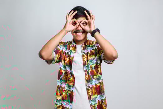 Man wearing casual shirt doing ok gesture like binoculars sticking tongue out, eyes looking through fingers photo