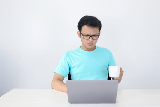 Young Asian man is serious when work in laptop with drink coffee in white glass. Indonesia Man wear blue shirt Isolated grey background. photo