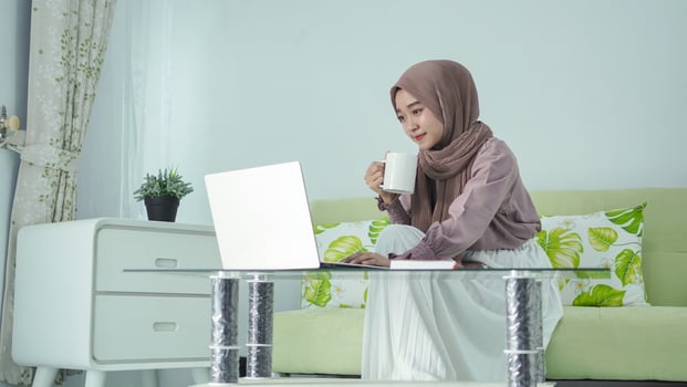 asian woman in hijab working from home using laptop while enjoying a drink photo