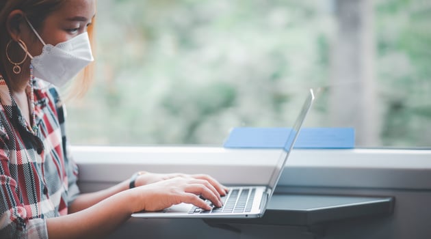 Businesswoman wearing a mask and working on laptop on a train, Asian freelance writer or designer writing emails on computer, working and smile resting on comfortable armchair near window of train. photo