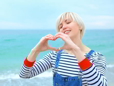 Beautiful young blonde caucasian woman with hands as love sign on vacation. Woman in striped blouse, sneakers and denim overall on the beach by the amazing blue sea background. Happy to be at the sea photo