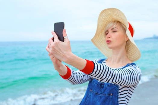 beautiful young blonde caucasian woman on vacation in straw hat, striped blouse nad denim overalls making selfie on smartphone by the amazing blue sea background photo