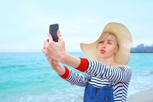 beautiful young blonde caucasian woman on vacation in straw hat, striped blouse nad denim overalls making selfie on smartphone by the amazing blue sea background photo