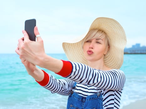 beautiful young blonde caucasian woman on vacation in straw hat, striped blouse nad denim overalls making selfie on smartphone by the amazing blue sea background photo