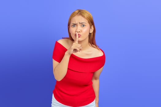 Portrait of kind Asian woman with short brown hair in red dress holding index finger on lips while standing isolated over purple background photo