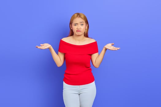 Portrait of attractive confused young Asian woman with short blonde hair in red dress standing and shrugging shoulders isolated over purple background photo