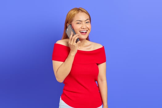 Portrait of happy young Asian woman in red dress rejoicing while talking on mobile phone isolated on purple background photo