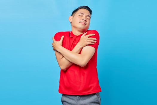 Portrait of cheerful asian young man in red shirt hugging himself and feeling cold isolated on blue background photo