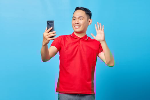 Portrait of cheerful Asian young man making video call and waving at smartphone camera isolated on blue background photo