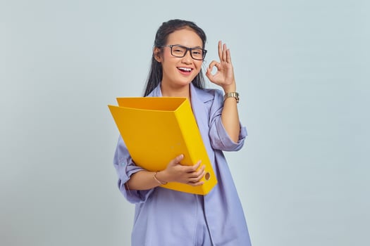 Portrait of cheerful young Asian business woman holding document folder and showing ok gesture isolated on purple background photo
