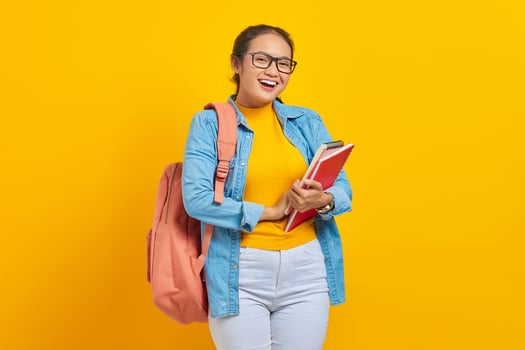 Portrait of cheerful young Asian woman student in casual clothes with backpack holding book and looking at camera isolated on yellow background photo