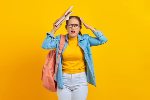 Dizzy young woman student in denim clothes with backpack, putting book and hands on head isolated on yellow background. Education in high school university college concept photo