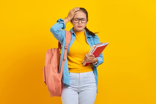 Beautiful young Asian college student in denim outfit with backpack holding notebook and looking dizzy with headache on yellow background. Education in high school university college concept photo