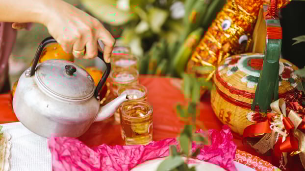 Person's hand is holding stainless steel teapot to pour tea into mug that is set on a table covered with red cloth for good fortune according to Chinese beliefs. photo