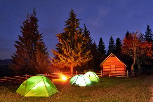 Night camping. Tourist have a rest at a campfire near illuminated tent and wooden house under amazing night sky full of stars and milky way. Astrophotography photo