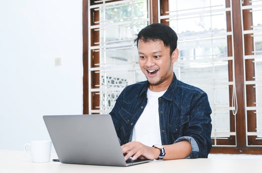 Young Asian man feeling happy and smile when work laptop on table. Indonesian man wearing blue shirt. photo