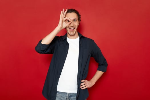 agitated man brings his hand to his eye showing ok sign. dressed in casual clothes. isolated on red background with space for advertising text. concept - people, body language, luck, victory, planning photo