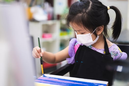 Portrait cute kid girl wearing mask while studying art in class. Children are working on painting crafts. Child wearing black apron is 5 years old. photo