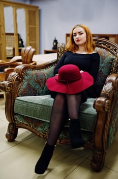 Fashion red haired girl with red hat and wear on black dress with bright make up sitting on royal armchair. photo