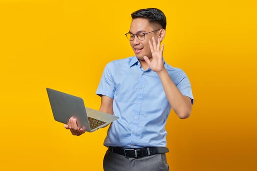 Portrait of cheerful young asian man Asian in glasses using a laptop, doing a video call, gesturing hi to friends isolated on yellow background. businessman and entrepreneur concept photo