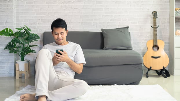 Smiling asian man wearing casual clothes sitting on a couch at the living room. Asian man using mobile phone chatting in living room. photo