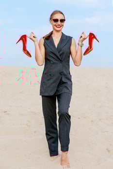portrait of stylish white young woman in suit, sunglasses, standing with the glossy high heels shoes in her hands outdoor photo