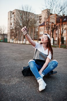 Young teenage urban girl with skateboard, wear on glasses, cap and ripped jeans at the yard sports ground on sunset making selfie on phone. photo