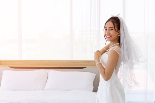 An Asian bride dressed in a white wedding dress stands in a cute and bright smile beside the bed in the room. photo