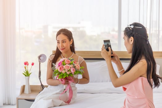 The bride's friend is using a smartphone to take a photo of an Asian bride in a wedding dress in her hand, holding a large, beautiful bouquet of flowers sitting on the bed.