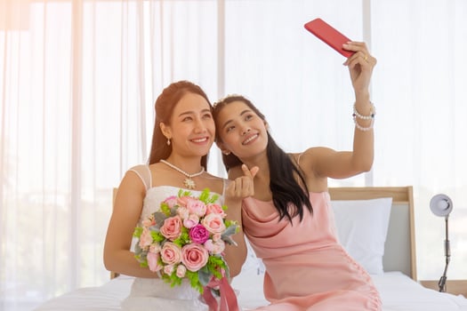 The bride's friend is using a smartphone to take a selfie of an Asian bride holding a bouquet of beautiful flowers in a wedding dress in the dressing room on the bed and making a mini heart hand sign. photo