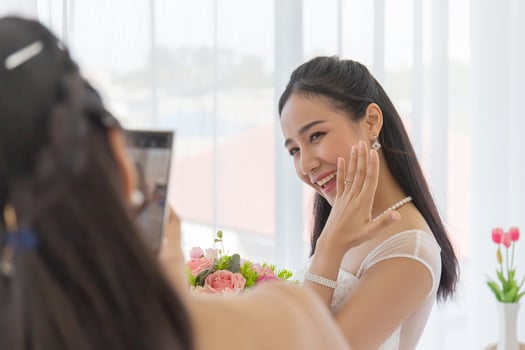 The bride's friend is using a smartphone to take pictures of an Asian bride in a wedding dress that shows her left-hand diamond ring in the dressing room with a cute, bright smile. photo