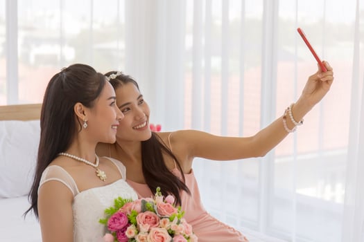 The bride's friend is using a smartphone to take a selfie of an Asian bride holding a bouquet of beautiful flowers in a wedding dress in the dressing room on the bed. photo