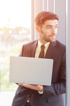 A business man dressed in a neatly dressed suit holds a tablet next to a glass wall in his office with a bright smile and a beautiful orange light. photo
