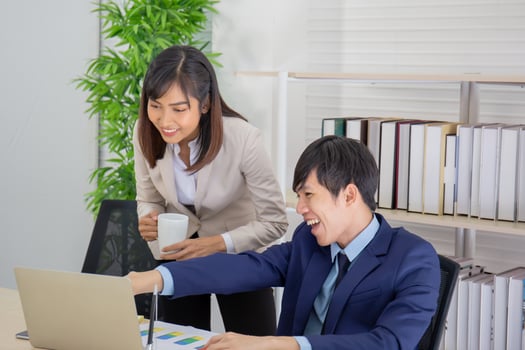 An Asian female coworker stood talking to a business man on the desk in her hand holding a cup of coffee. photo