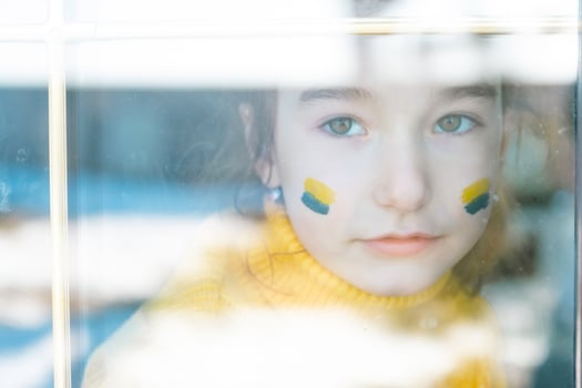 A sad child look at the window with the flag of Ukraine painted on the cheek, worries and fear. Humanitarian aid to children, world peace, security. photo