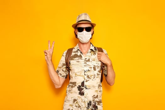 young male tourist traveler with backpack in a hat, sunglasses, a medical mask shows a peace sign with his hand isolated on a yellow background. the concept is recreation, people, adventure, vacation. photo