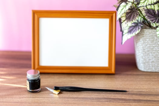Blank landscape wooden frame beside a small houseplant and calligraphy tools, painting or artwork display mockup in front of pink wall photo