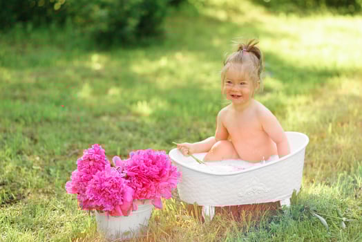 Happy toddler girl takes a milk bath with petals. Little girl in a milk bath on a green background. Bouquets of pink peonies. Baby bathing. Hygiene and care for young children. photo