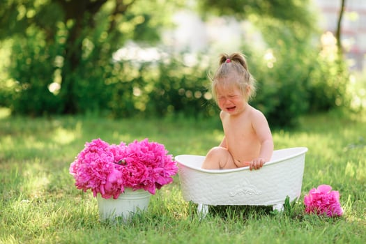 Happy toddler girl takes a milk bath with petals. Little girl in a milk bath on a green background. Bouquets of pink peonies. Baby bathing. Hygiene and care for young children. photo