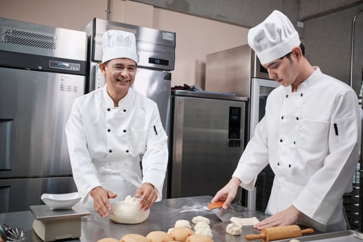 Two professional Asian male chefs in white cook uniforms and aprons are kneading pastry dough and eggs, preparing bread and fresh bakery food, baking in oven at stainless steel kitchen of restaurant. photo