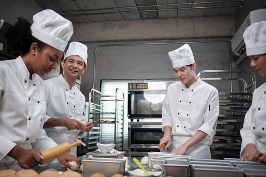 Multiracial professional gourmet team, four chefs in white cook uniforms and aprons knead pastry dough and flour, prepare bread, and bakery food, baking in oven at stainless steel restaurant kitchen. photo