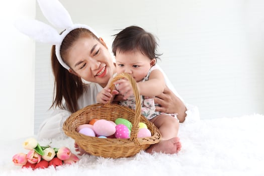 Happy smiling adorable six months baby girl playing with wicker basket of colorful Easter eggs in embrace of mother arms, mom with rabbit ears headband holding her sweet little daughter kid. photo