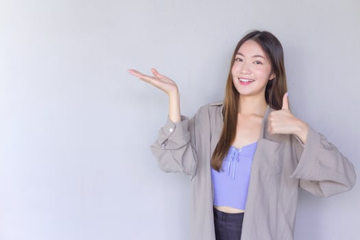 Beautiful young Asian woman wearing a purple shirt is acting thumb up as a good symbol and another hand shows as presenting something on the background. photo