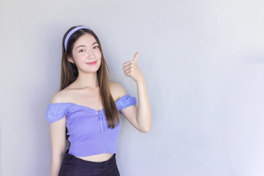 Young beautiful Asian woman wearing a purple shirt is acting thumb up as a good symbol and another hand shows as presenting something on the background. photo