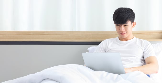 Young freelance Asian man working from home using laptop computer while sitting on the bed with copy space photo