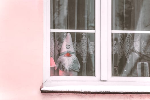 Felted toy gnome standing behind window with white tulle curtain in pink wall building photo